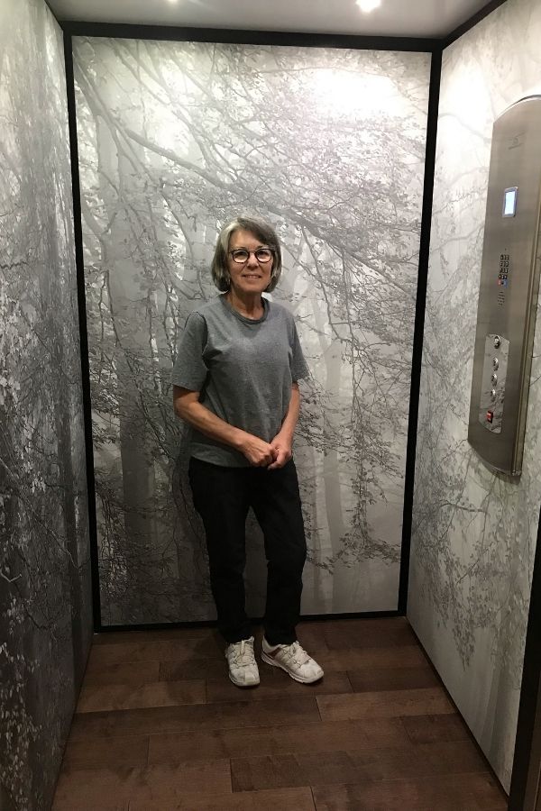 Autumn is Here Black and White Wall Mural, as seen behind this customer in her elevator, is a gray forest wallpaper from About Murals.