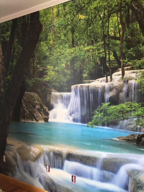 Erawan Waterfalls Wall Mural, as seen on the wall of this room, is a photo mural of blue water trickling over rocks in a Thai forest from About Murals.