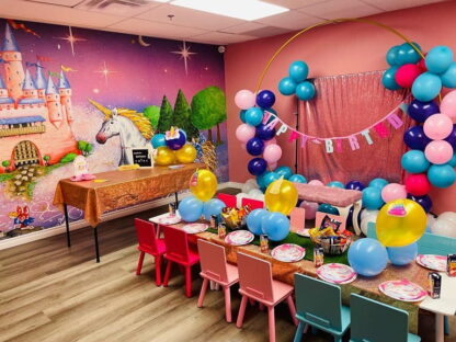 Princess Castle Wallpaper, as seen on the wall of this birthday party venue, is a girls wall mural of a pink castle, which unicorn and green frog prince on a purple background from About Murals.