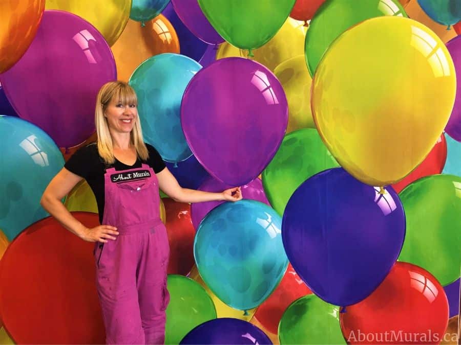 Adrienne of AboutMurals.ca stands next to a party room wallpaper full of colourful balloons