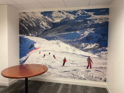 Whistler Ski Wallpaper, as seen on the wall of this office, is a photo mural of skiiers and snowboarders coming down the side of a snow covered mountain from About Murals.